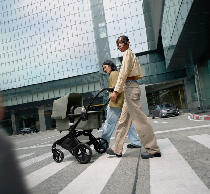 Ein stilvolles Paar spaziert selbstbewusst mit seinem Neugeborenen in einem Bugaboo Fox 5 Kinderwagen. Sie sind von gläsernen Wolkenkratzern umgeben.