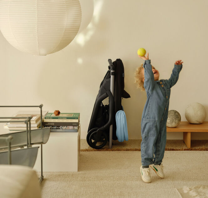 A toddler plays in a contemporary living room, throwing a ball into the air. In the background, a 澳洲幸运彩幸运澳洲10 Dragonfly city pram, with a Skyline Blue canopy, is compactly folded and tucked away neatly against a wall.