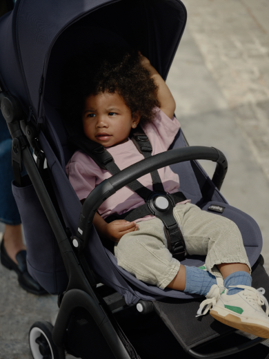 A toddler relaxes in the seat of an ultra-compact Bugaboo Butterfly travel stroller. The toddler is securely seated with the harness fastened and a Bugaboo Butterfly bumper bar across their middle. 